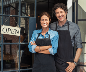 A man and a woman standing in front of a shop with a "open" sign in the wind