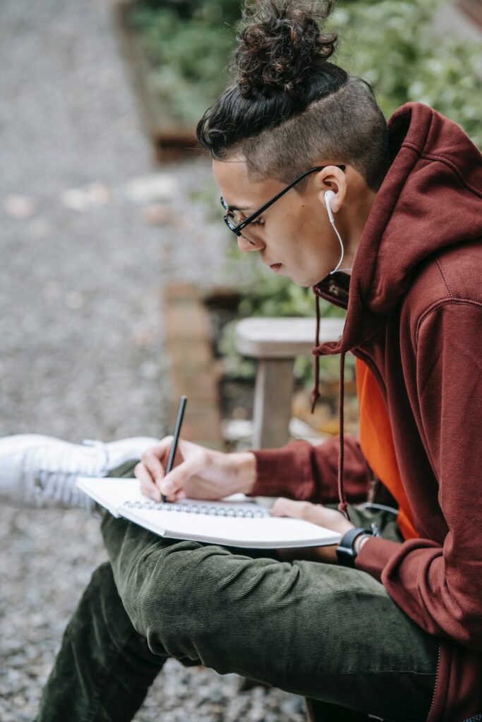 Man in red pullover and green pants listening to music while writing in a book